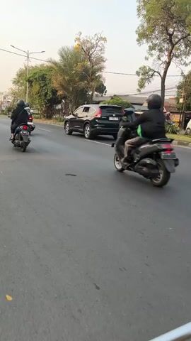 The video captures a street scene with several vehicles. In the foreground, a person on a dark-colored scooter is riding on an asphalt road, heading away from the camera. The rider is wearing a black jacket and a helmet. To the left and further back, another person on a scooter is also riding away from the camera. This scooter appears to be black. In the middle ground, a dark SUV is parked on the side of the road. Beyond the vehicles, there are trees and some buildings visible, suggesting an urban or suburban environment. The lighting indicates it is daytime, possibly late afternoon due to the warm tones in the sky. The road appears to be multi-lane, and there is a sidewalk or grassy area to the left of the road.