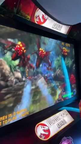 The video shows a man playing a Jurassic Park Arcade game with two young children. The game involves shooting at dinosaurs displayed on the screen. The arcade game is red with a large screen and a Jurassic Park Arcade logo. The man is operating the controls with one of the children helping him, while the other child looks on curiously. The environment appears to be an arcade.