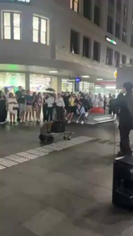 A street performer is rapping or beatboxing to a crowd in a city square at night. He is standing on a black speaker, holding a microphone. A keyboard is visible next to him. The audience is gathered in front of him, some sitting on metal pallets. There is a grocery store in the background, and streetlights illuminate the scene.  Some in the audience are holding umbrellas. There is a small cart with equipment on wheels in front of the performer.