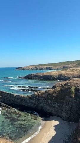 A vertical video captures a scenic coastal view under a clear blue sky. The foreground features rugged, dark grey rocks jutting out into the turquoise sea. The water is clear, revealing the sandy bottom in some areas and patches of seaweed. Small waves gently break against the rocks. Behind these, a small sandy beach is visible. In the mid-ground, more rocky outcrops and the rolling coastline extend into the distance. The landscape is covered in dry, golden grass and sparse vegetation. The horizon shows the endless expanse of the sea meeting the sky. The overall impression is one of natural beauty and tranquility.