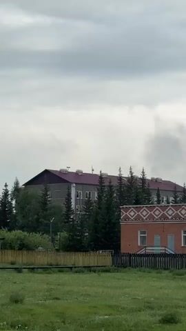 The video shows an exterior view of buildings and trees under a cloudy sky. In the foreground, there is a grassy field and a wooden fence. Behind the fence, there are two buildings. The building on the left is made of brick and has a decorative pattern around the top. The building on the right is larger and appears to be an apartment building with a maroon roof. Several evergreen trees are visible between and behind the buildings. In the distance, there is a tall, thin smokestack and another building with a gray roof. The sky is filled with gray clouds, with some patches of blue sky visible. The video pans slowly across the scene.