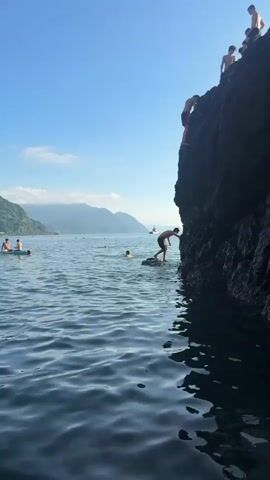 A person is seen jumping off a rock into the water. The water is blue and calm, with gentle waves. In the background, there is a beach with people sunbathing and swimming, and a coastline with green hills and mountains. The sky is clear and blue.