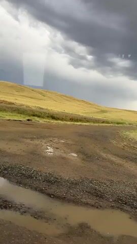 A dirt road is visible in the foreground with patches of water and gravel. Beyond the road is a grassy field that slopes upwards towards a hill. The sky is cloudy with dark storm clouds in the distance. The camera is moving forward, as indicated by the panning motion of the landscape. The overall scene depicts a rural landscape with signs of recent or ongoing rain.