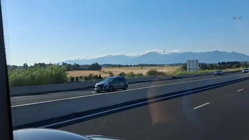 The video shows a highway scene from the perspective of a vehicle traveling on it. In the foreground, a dark blue SUV is visible in the right lane. In the distance, several other vehicles are on the highway, including a white car in the left lane and a white truck further ahead. The landscape to the left of the highway consists of fields and scattered trees. In the background, a mountain range is visible under a clear blue sky, with some clouds partially obscuring the peaks. The sun appears to be shining, casting long shadows. The highway has multiple lanes and a concrete barrier separating the lanes of oncoming traffic from the direction of travel.