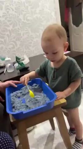A toddler with blond hair and fair skin stands at a small wooden table, playing with kinetic sand. He wears a green shirt and has on a diaper with striped socks. The kinetic sand is contained in a blue plastic tub. The toddler uses a yellow plastic spoon to scoop sand into a clear plastic cup. A person's hand, wearing a navy and white polka-dot shirt, rests on the edge of the table near the sand. The background features a gray and white patterned wallpaper, a dark brown dresser with various items on top, and a partially visible open white door.