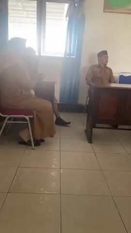 The video shows an indoor scene with several people. In the foreground, a person wearing a tan uniform and a headscarf is seated on a chair. To their right, another person in a similar tan uniform and a black cap is seated behind a wooden desk. Further to the right, another person in a white shirt and a headscarf is seated behind a desk. The room appears to be an office or meeting space, with tiled floors, desks, chairs, and a window with blue curtains. A bulletin board with text and graphics is visible on the wall. The lighting suggests daytime.