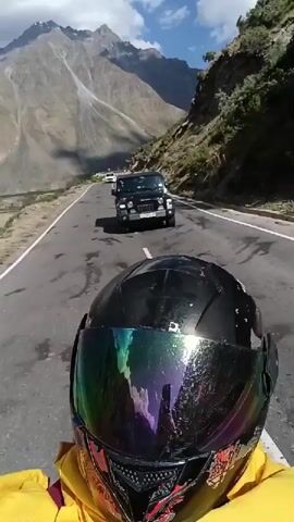 A first-person perspective video shows a motorcycle rider wearing a black helmet with a rainbow-tinted visor. The rider is on a paved road with a white line down the center. In front of the rider, there are two vehicles: a black SUV, possibly a Jeep, and a white car further ahead. The road is flanked by a steep, rocky mountain slope on the right and a more gradual, vegetated slope on the left. The background features large, barren mountains under a cloudy sky. The rider is wearing a yellow jacket.