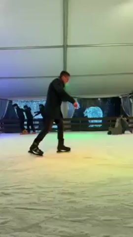 The video shows an indoor ice-skating rink with multiple people skating. The rink is inside a white tent-like structure with a white roof. Light blue and green lights illuminate the ice. In the background, there's a barrier around a decorative winter scene. Several people are seen ice skating at various levels of experience. One person uses a brown skate aid to help them balance. There are a few other people skating more confidently. The ice surface shows skate marks.