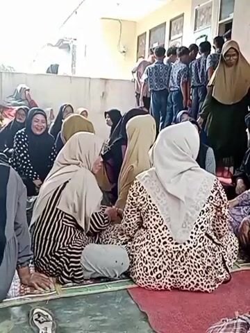 The video shows a gathering of people, mostly women wearing hijabs, seated on mats on the floor. In the background, a group of young people, likely male students in uniform, are standing. The environment appears to be an outdoor area or a covered patio with a building in the background. Some people are interacting with each other, with some of the women facing and speaking to one another. The overall atmosphere seems to be one of a community event or gathering.