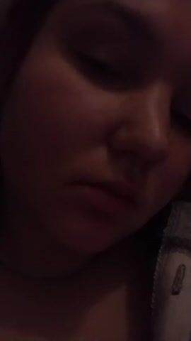 A person's face is shown close up, slightly angled down to the right. The person has light skin, with red undertones, and their eyes are looking down. Their lips purse slightly. They are wearing a gray and black checkered shirt. A silver chain is visible at the base of their neck. The lighting is dim.