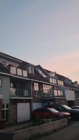 The video shows a residential street lined with houses and parked cars under a pink and blue dusk sky. The camera slowly pans from left to right, revealing more of the street and houses. There are various types of cars parked along the road, and the houses have different architectural styles. Some houses have balconies, while others have more traditional rooflines. There are also trees and bushes visible along the street. The sky has a soft, pastel hue with streaks of pink and blue, suggesting it is either sunrise or sunset.