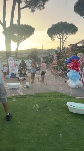 The video shows a daytime outdoor scene with a sunset backdrop and numerous bubbles floating in the air. In the foreground, a grassy area has a white bathtub with green liquid and a few scattered bubbles. In the middle ground, a paved area features several people, some of whom are interacting with bubble-making equipment. A red and white striped tent-like structure and clusters of red, white, and blue balloons are visible to the right. Several large trees are present in the background, and a hazy cityscape can be seen in the distance. The overall atmosphere appears to be festive, possibly a party or event, with a focus on bubble entertainment.