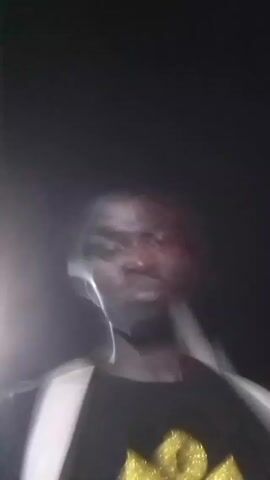 A man with dark skin is holding a phone and recording a video of himself at night. He is standing on a sidewalk next to a road with streetlights. He is wearing a black t-shirt with a gold design and a light-colored backpack. He makes various facial expressions, including sticking out his tongue and rubbing his chin. The background is blurry due to the low light and movement.