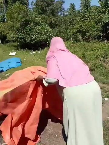 A person wearing a pink hijab and a light blue long-sleeved shirt and light green skirt is standing on a dirt ground. The person is interacting with a large orange tarp that is partially unrolled and spread out on the ground. The tarp has a lighter colored section in the middle. The background consists of green grass, trees, and some bushes under a clear blue sky. There are also some other colored tarps visible in the background, one blue and one white. The person appears to be moving or adjusting the orange tarp.