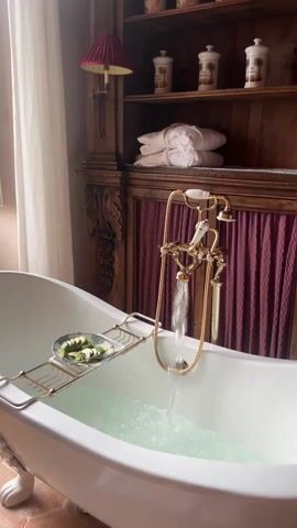 The video displays a luxurious bathroom. A vintage-style, white clawfoot bathtub is filling with water. The ornate gold-colored faucet is running. A rack spanning the tub holds a small plate of cucumber slices. In the background, a rustic wooden cabinet displays neatly folded white towels and decorative jars. Natural light streams in through a window with white shutters, revealing a green landscape with trees. A red lampshade is visible on the cabinet, adding to the room's warm aesthetic. The floor appears to be tiled.