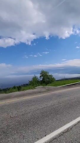 A vertical video shows a view from a roadside looking toward a body of water and distant mountains under a cloudy blue sky. The foreground features a section of asphalt road with a yellow dividing line and a white edge line. A metal guardrail runs parallel to the road, separating it from a grassy embankment. On the embankment, several green trees are visible. Beyond the embankment, a hazy, mist-covered landscape stretches toward a mountain range in the distance. The sky is filled with large, puffy white clouds against a bright blue background. As the video progresses, the camera appears to move slightly, causing minor shifts in the perspective of the scene. In the far distance, a solitary signpost with a rectangular board is visible on the right side of the frame. The overall impression is of a scenic view from a highway, likely in a mountainous or coastal region.
