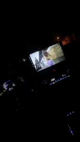 The interior of a car is seen at night. The interior is dark, but the dashboard gauges and infotainment screen are visible. A music video is playing on the infotainment screen, showing a man with colorful paint on his face.  The camera is stationary.  The only source of light besides the instrumentation is the infotainment screen.