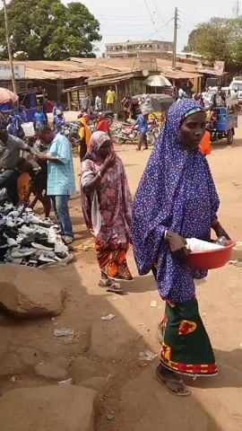 The video shows a busy marketplace in what appears to be a West African country. Several people are gathered around a pile of shoes, apparently for sale. People are inspecting the shoes. A woman in a colorful headscarf and dress walks by, followed by another woman in similar attire. There are small shops or stalls in the background with corrugated metal roofs. Motorcycles and other vehicles are parked nearby. The ground is dusty and littered with trash. The scene conveys a sense of bustling commercial activity.