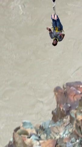 The video shows a person bungee jumping. The jumper is suspended by a cord attached to a harness, and is falling towards a muddy river below. The jump appears to be taking place from a high vantage point, possibly a bridge or cliff. The surrounding environment is mountainous and rugged. The jumper is wearing jeans and a dark colored shirt, and the bungee cord is white. The video is shot from a perspective that emphasizes the height and the danger of the activity.