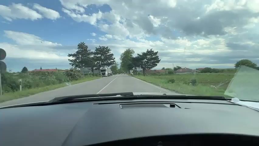 The scene is viewed from inside a car driving down a straight two-lane road. The car is approaching the entrance to a village. On the left side of the road, a white truck with a large, green trailer is stopped, partially blocking the lane. A person is standing near the truck, appearing to work on it. There are orange cones around the truck, indicating a potential hazard. On the right side of the road, there is a sign indicating the village name in Cyrillic. The sky is partly cloudy with blue patches. The surrounding area consists of fields and some houses.