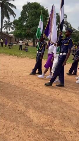 A small group of people, dressed in navy blue uniforms with white belts and caps, are marching on a dirt field. They are carrying three flags: one green and white, one white and black, and one white with an unknown design. The person carrying the white and black flag is in the foreground. A few other people are in the background, watching the procession. The field is surrounded by trees and buildings. The sky is overcast.