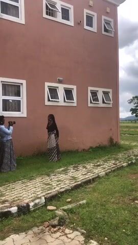 The video shows a person posing for a photograph against a tan-colored building. The person is wearing a black top and a patterned skirt. A second person is holding a camera and appears to be taking the photograph. The building has multiple windows, some of which are open. The ground is covered with grass and a paved walkway. The sky is cloudy.