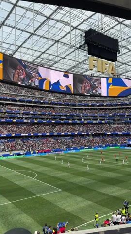 A wide shot captures a soccer match at a stadium. The stadium has a large, arched, translucent roof. A massive video screen above the field displays graphics and images, including the FIFA logo. The field is green with white markings, and players in red and white uniforms are spread across it. Spectators are seated in tiered stands surrounding the field, their faces obscured by distance. The atmosphere appears to be that of a live sporting event.