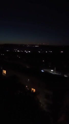 A high-angle shot captures a dark landscape dotted with distant lights. The foreground is almost entirely black, with faint outlines of buildings or structures visible in the lower left and center. These structures have illuminated windows, suggesting interiors. Further into the scene, a road or pathway is faintly visible, marked by white lights that could be streetlights or vehicle headlights. The horizon is marked by a faint glow of light against the deep blue of the night sky. The overall impression is of a city or town viewed from a distance at night.