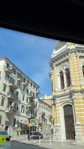 The video, taken from inside a moving vehicle, shows a street view in a European city on a sunny day. The camera is pointed upward, capturing the buildings lining the street and the clear blue sky. On the left are buildings with white facades, green shutters, and balconies with wrought iron railings. The Croatian flag is visible on one of the balconies. On the right is a yellow building with ornate architectural details. Traffic signs for pedestrian crossings and yielding to oncoming traffic are visible. A yellow taxi is driving on the left side of the road. The movement suggests the car is proceeding down the street, providing a dynamic view of the urban landscape. Pedestrians are visible walking in the distance.