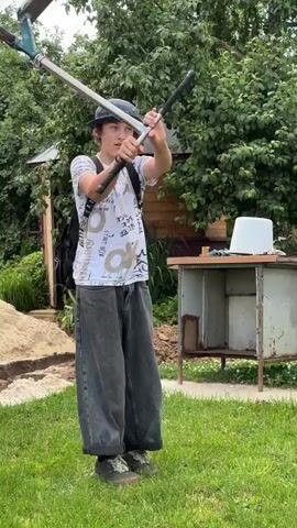 A young person with dark hair wears a blue cap, a white t-shirt with graphics, and baggy dark gray pants. They have a black backpack on and are holding a scooter with both hands, appearing to be in the process of performing a trick. The scooter has a green and white deck and black handlebars. The person is standing on a green grassy lawn in what appears to be a backyard. Behind them, there is a sand pile on the left, a rustic metal table with a white bucket on it, and lush green trees and foliage in the background. The sky is overcast.