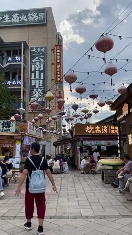 A narrow pedestrian street is lined with shops and stalls, decorated with numerous red and yellow lanterns and string lights. The ground is paved with light-colored tiles. In the foreground, a man wearing a black t-shirt, red pants, and a light blue backpack walks away from the camera. To his right, a woman in a patterned blouse and dark pants walks towards him, carrying a bag. They appear to be interacting, possibly engaged in conversation. Further down the street, more people are seen sitting at tables outside shops and walking along the pathway. The buildings on either side of the street have multiple stories, with signs in Chinese characters indicating businesses such as a hot pot restaurant. The sky above is partly cloudy.