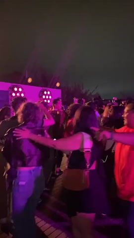 A group of people are dancing at an outdoor event at night. They are illuminated by pink and purple stage lights. The individuals are dressed in casual attire. Some are smiling and appear to be enjoying themselves. The background is dark, suggesting it is nighttime. There are multiple beams of light cutting through the darkness from above, indicating the presence of stage lighting equipment. Some individuals are holding drinks. The scene depicts a lively social gathering.