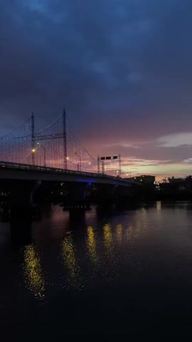This is a vertical video showing a bridge at dusk. The sky is dark blue with purple and pink hues of sunset visible on the horizon. The bridge is illuminated by strings of lights that follow the suspension cables, creating a festive appearance. Lights are also visible on the bridge's roadway and reflected in the dark water below, which appears to be a river. The water's surface is slightly rippled, distorting the reflections. The overall scene is serene and atmospheric, capturing the transition from day to night. There are no people visible in the video.
