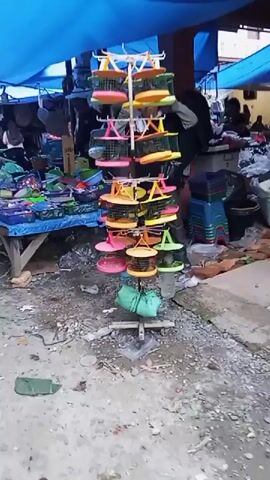 The video shows a close-up view of a revolving display of colorful plastic objects, possibly bird feeders or food dishes, at an outdoor market. The objects are arranged in tiers on a vertical stand. The background is blurred but appears to show more stalls with various items, including more plastic containers, and a few people walking by. The ground is uneven and appears to be dirt or gravel. The overall impression is a busy, informal marketplace.