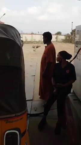 The video shows a CNG (Compressed Natural Gas) station. There are two men and one woman visible. One man in an orange robe stands next to the gas pump while the woman, wearing a black uniform with a red logo, sits on a red barrier nearby. The gas pump is stainless steel. A white truck with "CNG" written on it is parked at the pump. Behind a green pillar with a "Caution C.N.G. Area" sign is another man in a plaid shirt and dark pants. There is a small, wooden table nearby, cluttered with papers. The sky is overcast.