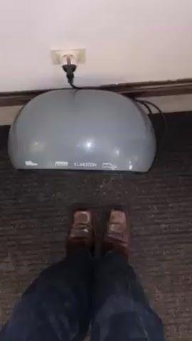 The video shows a person standing in front of a gray Klarstein shoe dryer. The person is wearing brown dress shoes and blue jeans. The shoe dryer is plugged into a white electrical outlet in the wall. The shoe dryer is a gray, dome-shaped object with the word "KLARSTEIN" printed on the side along with the image of shoes. The person pushes down on the dryer with their foot. A motor whirs to life and it begins to function. The floor has gray carpet.