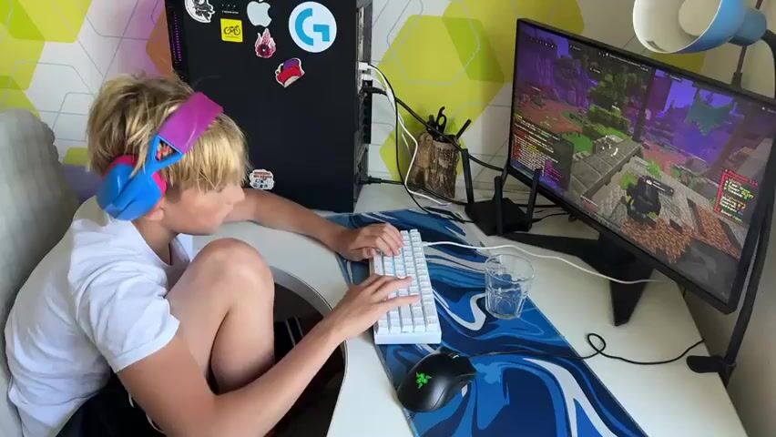 A person with blond hair, wearing a white t-shirt and black shorts with a striped pattern, is seated at a desk. They have large, colorful headphones on, with pink and blue earcups. Their head is bowed, and their hands are on a white keyboard. A computer monitor displays a colorful, pixelated game. To the right of the keyboard is a glass of water and a black gaming mouse with a green logo. Behind the monitor, a black computer tower is visible, adorned with stickers, including a blue G logo. The desk has a blue and white swirled mousepad. The background features a wall with a geometric pattern of green and yellow shapes. The person appears to be in the process of playing the game.