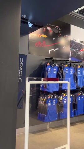The video shows the inside of a Formula 1 racing-themed store. The first shot shows a rack of Red Bull Racing apparel beneath racing posters. The camera then pans across a wide selection of clothing. A Formula 1 race car is positioned in the center of the store. Several people browse the merchandise.