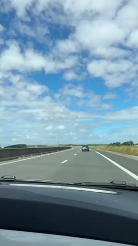 The video shows a point-of-view shot from inside a car driving on a highway. The road has multiple lanes with white markings. The sky is blue with scattered white clouds. On the right side of the road, there is a guardrail and fields with some trees in the distance. There are speed limit signs visible. A dark-colored car is ahead in the same lane. The dashboard and windshield wipers are visible in the foreground.