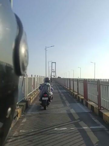 A motorcycle is traveling across a bridge. The motorcycle is carrying two people. The person in front appears to be driving the motorcycle, while the person in the back is wearing a backpack. The bridge has a sidewalk or pedestrian lane separated from the road by a fence. There are lampposts along the bridge and a large suspension bridge structure is visible in the distance. The sky is clear and the sun is shining brightly. The video is shot from the perspective of someone on another motorcycle.