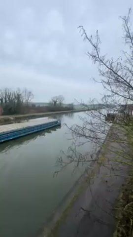 The video shows a canal on a cloudy day. A long, flat, blue barge is traveling down the canal, leaving a wake in the water. The canal is bordered by grassy banks with trees and some buildings in the distance. A red metal structure, likely part of a bridge, is visible in the foreground, featuring numerous rivets. There's no human presence readily apparent in the video frame.