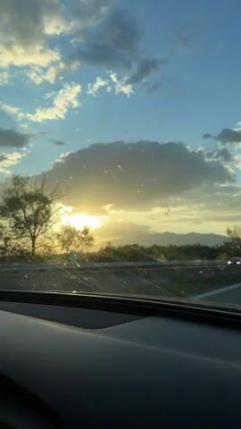 The video shows a sunset seen from inside a car, likely from the dashboard. The sky is blue with scattered clouds, and the sun is partially obscured by a larger cloud, creating a bright, golden effect. There are power lines and trees on either side of the road. Cars and a semi-truck pass in the distance. The windshield is dusty and scratched.