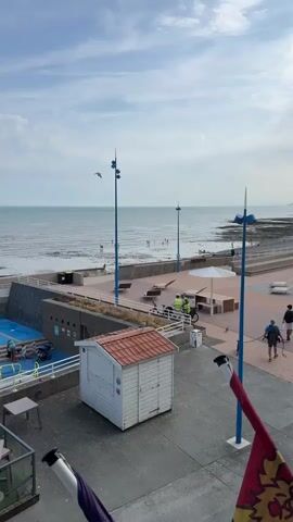 The video begins with a wide shot of a beach promenade. The sky is cloudy with a few patches of blue. The sea is calm, with small waves. In the foreground, there is a small white shed with a red roof. A seagull is perched on the roof. Further back, a paved promenade runs along the coastline. There are several lounge chairs and benches along the promenade. Two people in yellow safety vests are standing near bicycles. In the distance, several people are visible on the beach and in the water. The video zooms in slightly to focus on the seagull and the people on the promenade. The overall atmosphere is relaxed and peaceful.