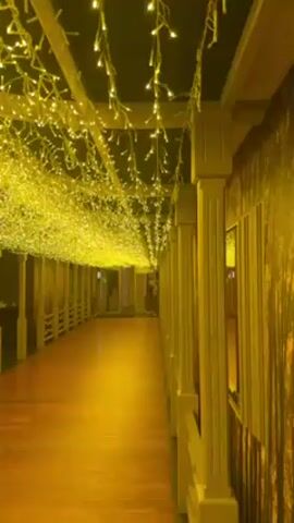 A static shot shows a well-lit hallway adorned with numerous yellow string lights overhead. The lights create a dense, glowing canopy effect. The hallway has a wooden floor, and the walls are decorated with a vertical forest-themed pattern. On the right side of the hallway, there are white pillars connected by a white railing. A person wearing a white jacket and dark pants walks away from the camera down the hallway. The overall atmosphere is warm and inviting due to the color and arrangement of the lighting.