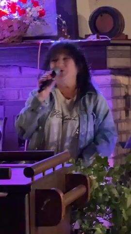 A person with dark hair and a denim jacket is holding a microphone and singing into it. They are standing behind a wooden railing and some greenery. In the background, there is a brick wall, some flowers, and a barrel. The lighting appears to be purple and blue. The person's mouth is open as they sing, and they are looking towards the microphone. Their right hand is extended towards the microphone, and their left hand is at their side.