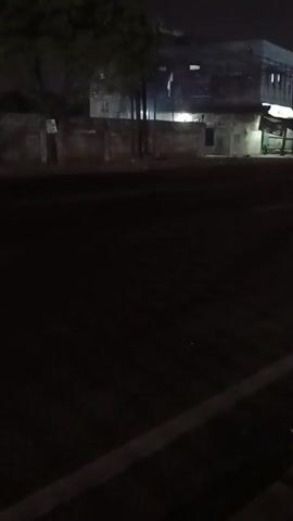 The video appears to be shot at night, likely from a moving vehicle. It shows a dark, paved road with a concrete curb in the foreground. In the background, there is a brick wall and a building with some lights on. There are also some trees visible. The scene is dimly lit, and the image is slightly blurry due to the low light conditions and potential movement. No people or specific actions are clearly discernible. The overall impression is of a quiet, urban or suburban street at night.