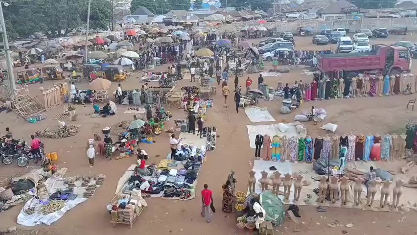 The video shows an outdoor market with many people browsing goods displayed on blankets, tables, and hangers. Vendors sit or stand by their merchandise, which includes clothing, produce, and various other items. Several motorcycles and vehicles are parked around the periphery of the market. The area is an open, dirt field with a few scattered buildings and trees in the background. The sky is overcast, casting a soft light over the scene.