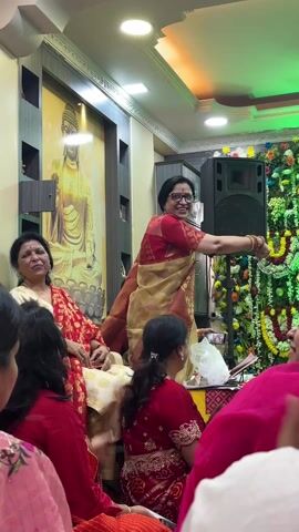 The video shows a group of people gathered in a room that appears to be set up for a celebration or religious event. Most of the people, predominantly women, are seated on the floor or on low seating. Some are dressed in traditional Indian clothing. One woman in a gold and red sari is standing and seems to be directing or overseeing the proceedings. Behind her is a speaker and a decorated backdrop, possibly a floral arrangement with lights. In the background, a large image or painting of a figure, possibly Buddha, can be seen on the wall. The room has decorative lighting with a green hue, suggesting it's part of the celebratory atmosphere.