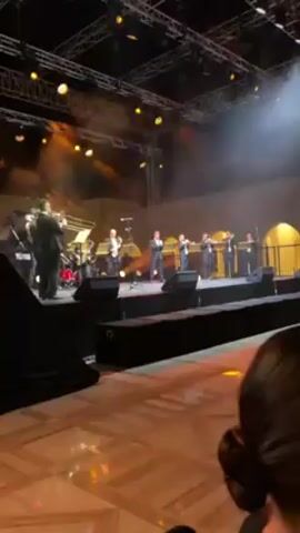 A medium shot shows a musical performance on a stage, viewed from the audience. The stage is elevated and lit by bright spotlights. A full band is on stage, including musicians playing brass instruments (trumpets, trombones, and a saxophone), as well as drums. The performers are dressed in dark suits. The stage backdrop is illuminated with orange light and some smoke. A person with dark hair is visible in the bottom right corner of the frame, likely part of the audience. The floor is patterned with a light-colored design. The overall atmosphere suggests a formal event.