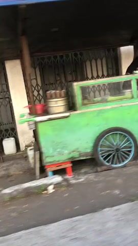 A blurry video shows a green food cart with a blue-wheeled cart parked on the side of a street. A metal accordion gate is visible behind the cart, as is a white plastic container. The cart appears to be unattended.